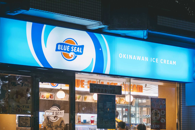 A vivid blue backlit opaline plexiglass lightbox sign for 'Blue Seal Okinawan Ice Cream' mounted above a shop entrance, glowing brightly against the night sky. This custom-printed opaline plexiglass lightbox panel (3 or 5mm thick, double-sided print) is ideal for shops, restaurants and commercial businesses seeking eye-catching illuminated signs that boost street visibility, attract customers and…