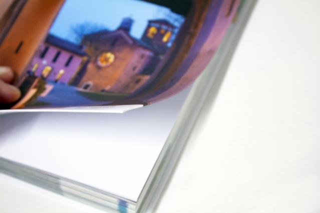 A close-up of a Dutch-binding flexible hardcover book being gently opened, revealing the foldable cover with a full-bleed evening photograph of an illuminated Italian piazza and church. The flexible cover of Dutch-bound books folds back completely, making them light, durable and easy to handle — ideal for travel guides, art books, local history publications and corporate catalogues.