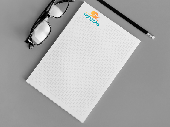 A custom A4 notepad with a small grid template and Horizons brand logo at the top, displayed with glasses and a pencil on a grey desk surface. Custom A4 notepads with printed templates — millimetred, dotted, squared or lined — are practical and appreciated promotional tools for companies and professionals who want to distribute a useful branded stationery item.