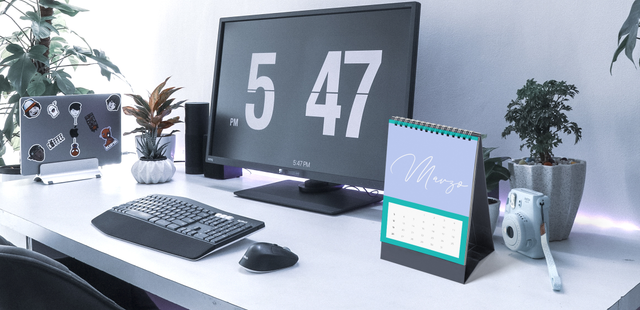 Compact spiral-bound desk calendar with custom cover, sitting on a modern workspace next to keyboard, monitor and plants. Shows how a small personalized calendar fits naturally on any office desk. Ideal as a corporate gift that stays in front of customers all year.