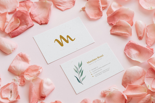 Two elegant white business cards photographed among pink rose petals — one showing a gold foil cursive 'M' logo, the other 'Massimo Rossi' with a delicate green botanical branch illustration. Personalised business cards for gardeners and florists with nature-inspired botanical motifs and gold foil accents project a premium, trustworthy professional image.