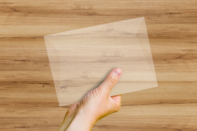 A semi-translucent GSK transparent envelope made of natural Fedrigoni paper held in a hand over a wooden surface, the wood grain visible through the paper. Sprint24 prints transparent GSK envelopes in natural translucent paper with no chemical agents — ideal for refined wedding invitations, brochures and content that benefits from a touch of see-through elegance.