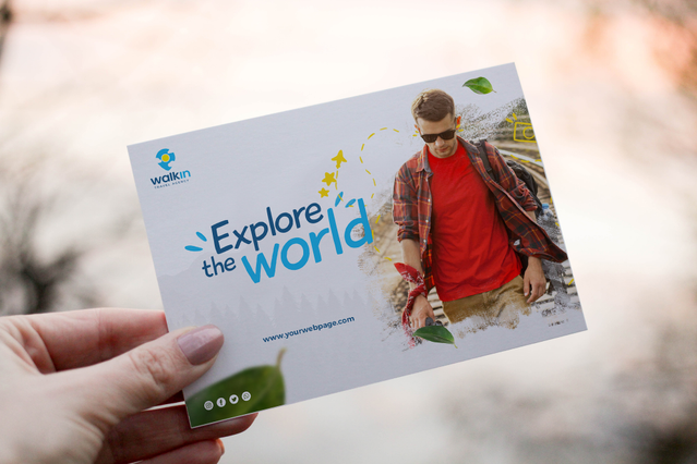 A hand holding a horizontal personalised postcard for the travel brand 'Walkin' featuring bold 'Explore the World' typography, a logo and a full-colour photo of a young man in sunglasses and red shirt. Custom postcards are a powerful and versatile marketing tool — ideal for travel agencies, event promotions, discount campaigns and direct mail advertising with high visual impact.