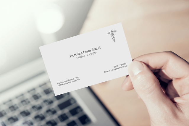A hand holding a clean white business card for 'Dott.ssa Flora Accuri, Medico Chirurgo' featuring a caduceus medical symbol logo, displayed near a laptop keyboard. Custom-printed medical business cards for doctors and surgeons help build professional credibility and patient trust — fully personalised with name, specialty, contact details and medical logo.