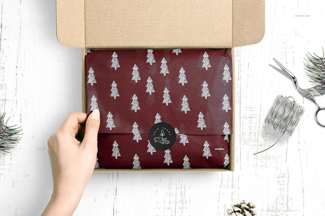 A hand opening a kraft cardboard box to reveal custom-printed burgundy wrapping paper with a silver Christmas tree pattern and a branded wax seal, surrounded by festive pine sprigs and ribbon. This personalised custom wrapping paper is ideal for boutiques, jewellers, confectioneries and artisan businesses seeking premium branded packaging that turns every product presentation into an elegant, mem…