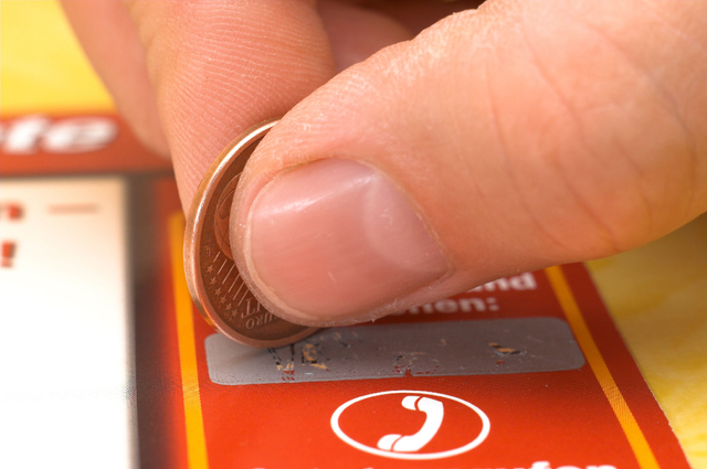 A close-up of fingers scratching the silver coating off a multiple-choice scratch card with a coin, revealing the answer area on an orange-printed card. Sprint24 prints multiple-choice scratch cards with up to 6 different answers under the silver patina — perfect for advertising campaigns, contests and customer engagement promotions.