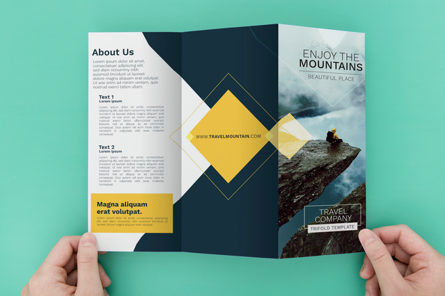 Two hands holding an open tri-fold brochure for a 'Travel Company' with 'Enjoy the Mountains' headline, a dramatic mountain climber photograph, gold diamond graphic accents and an 'About Us' text panel. Custom-printed tri-fold brochures with striking travel photography and bold typography are essential marketing tools for travel agencies, tour operators and adventure tourism brands.