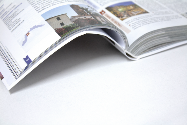 An open Dutch-binding flexible hardcover book showing a multi-column spread with travel photography, colour photos of buildings and landmarks, and editorial text columns, displayed on a white surface. Books with Dutch binding (brossura olandese) combine the durability of a hardcover with the flexibility and lightness of a softcover — ideal for travel guides, manuals, corporate books and children'…