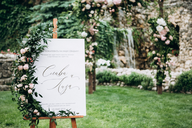 A white calligraphy wedding tableau mounted on a wooden easel against a lush floral arch backdrop in an outdoor ceremony setting. Sprint24 prints custom wedding tableaus in various materials and formats — ideal for organizing seating arrangements at wedding receptions with elegant, personalized name and table displays.