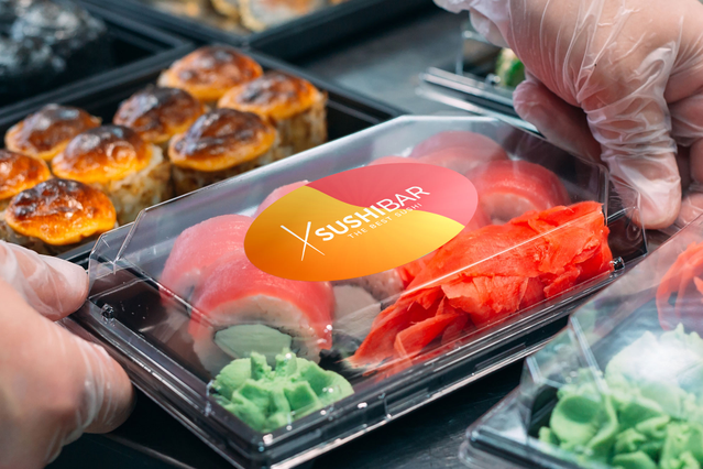 A bright orange oval custom vinyl sticker with a sushi brand logo applied to a transparent takeaway food container. The sticker stands out vividly against the colorful sushi contents and gloved hands preparing the order. Sprint24 custom vinyl stickers in oval shapes are perfect for food packaging, branding, and promotional labeling.