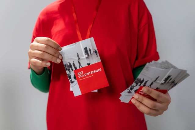 A person wearing a red shirt holds a stack of custom promotional postcards with bold red and white design featuring 'Art Volunteering' event branding. The scene illustrates how postcards are easily distributed at fairs, events, and public gatherings. Print promotional postcards online with Sprint24.