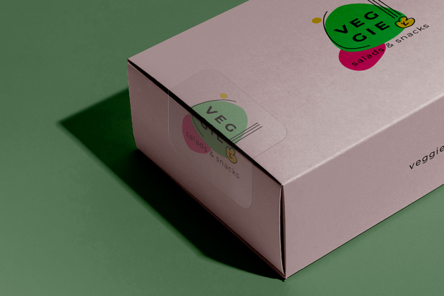 A pink rectangular product box decorated with a colourful 'Veggie' transparent PVC sticker featuring green and pink graphic dots. Sprint24 prints clear PVC stickers without white backing — ideal for product packaging, brand decoration on coloured surfaces and shop displays where the sticker integrates seamlessly with the design.