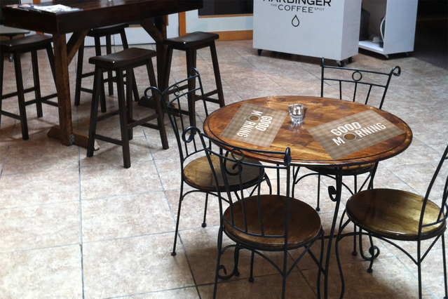 Custom printed placemat displayed on a wooden table in a modern cafe setting with black metal seating. Shows professional print quality suitable for branded table linens in restaurants and cafes. Personalized placemats enhance dining ambiance and hospitality branding.