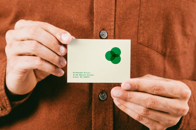 An eco-friendly natural recycled paper business card with a stylised green dot logo and contact details, held in both hands in front of an orange shirt. Sprint24 prints recycled paper business cards 100% biodegradable and free of plastic — ideal for eco-conscious brands, restaurants, clubs and professionals who value sustainability and originality.