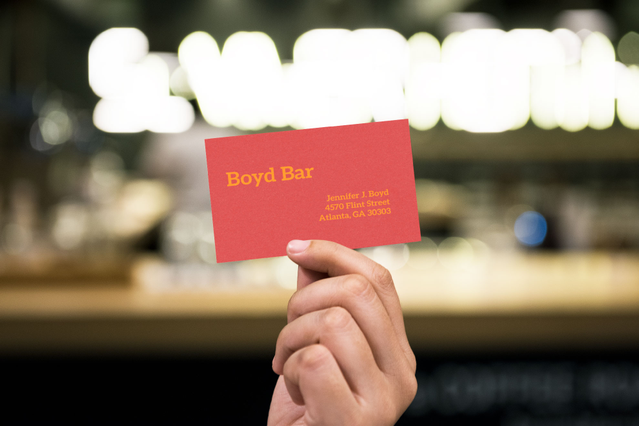 A bright red 'Boyd Bar' business card printed on coloured Sirio Fedrigoni paper with yellow text, held in front of a softly lit bar background. Sprint24 prints colour business cards in 6 vibrant Sirio paper shades with optional hot foil stamping — perfect for bars, restaurants and creative brands wanting bold, distinctive corporate identity.