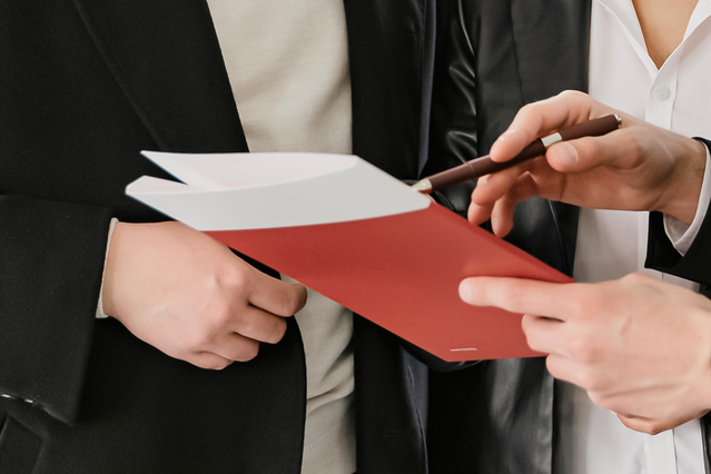 A vibrant red custom document folder being exchanged between professionals in a corporate setting, exemplifying how branded folders enhance business presentations and client impressions. Sprint24's customizable document folders feature pockets and flaps to organize materials while showcasing your company's identity during important meetings and events.