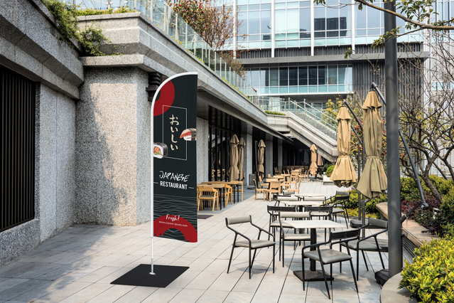 A tall custom-printed blade flag display standing on a terrace outside a restaurant, with branded print visible against an urban backdrop of outdoor seating. These personalised blade flags (4 available heights, water or steel base, indoor/outdoor use) are ideal for restaurants, shops and events seeking eye-catching outdoor advertising displays that boost street visibility, attract foot traffic an…