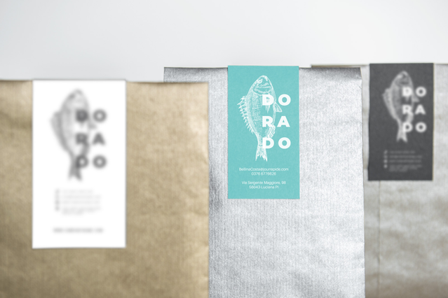 Three different paper bags — white, kraft and grey — each sealed with a custom-printed 'Dorado' fish-logo closure label in matching colourways: white, teal and dark charcoal. These personalised closure labels demonstrate how consistent branding across multiple colourways creates a cohesive premium packaging identity — ideal for food businesses, fishmongers and artisan producers seeking branded ad…