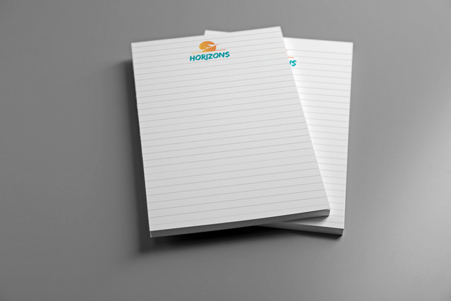 Two custom A4 notepads with a lined template and colourful Horizons travel brand logo at the top, displayed on a grey surface. Custom A4 notepads with lined, squared, dotted or millimetre templates and company branding are ideal promotional stationery items for travel agencies, companies and professionals to distribute at meetings, events and corporate gifting occasions.