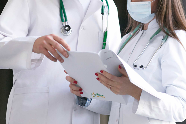 Two doctors in white coats with green stethoscopes reviewing a branded medical report folder together in a clinical corridor. Sprint24 prints custom medical report folders with die-cut pockets and clinic logos for doctors, hospitals and private practices — ideal for delivering patient exam results in professional, recognizable branded folders.