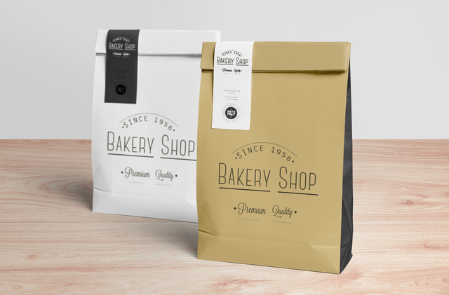 A white and a kraft paper bag for 'Bakery Shop — Since 1956 — Premium Quality' both sealed with custom-printed PVC closure labels showing consistent branding on a wooden surface. These personalised closure labels (square or shaped, various cut types, PVC material) are ideal for bakeries, food shops and retail businesses seeking branded adhesive seals that close packaging elegantly while keeping t…