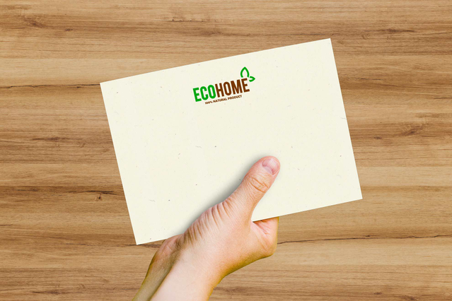A Woodstock Betulla envelope in light wood-tone recycled paper with a green 'ECOHOME' branded logo, held in a hand over a wooden surface. Sprint24 prints Woodstock Betulla envelopes in 80% recycled paper and 20% pure ECF cellulose — ideal for eco-conscious brands, sustainable communications and warm, natural-looking invitations and corporate mailings.